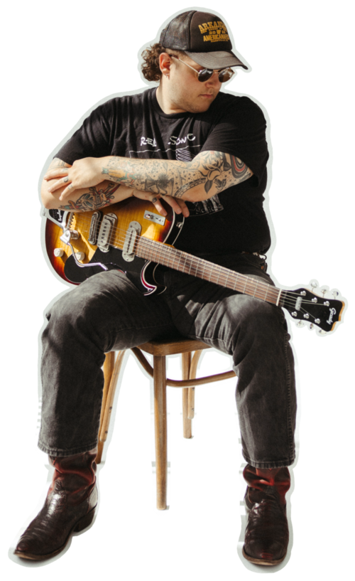 This is a cut-out photo of Grady Drugg sitting on a rattan chair and holding a sunburst-colored guitar. They are wearing a brown and gold trucker hat, golden wire-rim sunglasses, a black graphic t-shirt, black jeans, and black cowboy boots.